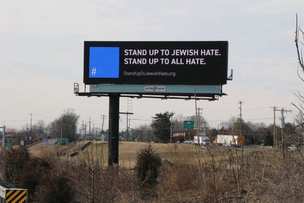 A large digital billboard on the side of a highway displays the message "STAND UP TO JEWISH HATE. STAND UP TO ALL HATE." along with a blue square symbol and the website StandUpToJewishHate.org.